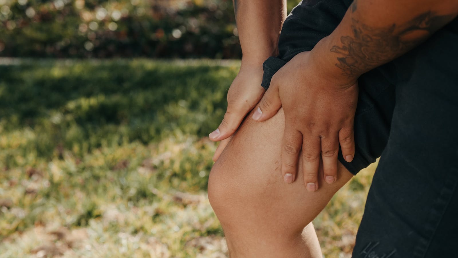 A close-up image of a person holding their knee, indicating pain or injury, with greenery in the background.